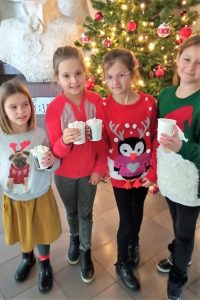 Cztery dziewczynki ubrane w świąteczne sweterki pozują z kubkami gorącej czekolady, w tle za nimi choinka.