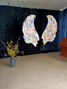 3.	Zdjęcie przedstawia scenę, na której widnieją białe skrzydła z kolorowymi piórami oraz szary wazon z gałązkami.