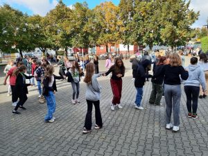 Uczniowie znajdują się na boisku szkolnym i tańczą belgijkę.
