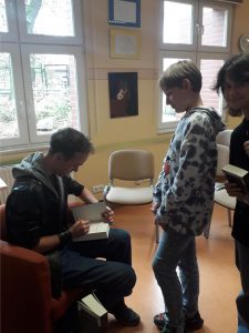 Młody chłopak siedzący w fotelu podpisuje książkę.  Przed nim dwoje uczniów.