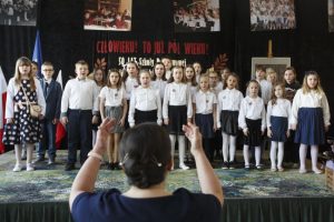 Szkolny zespół wokalny w trakcie występu - dzieci na scenie w strojach galowych; na 1. planie dyrygująca nauczycielka muzyki