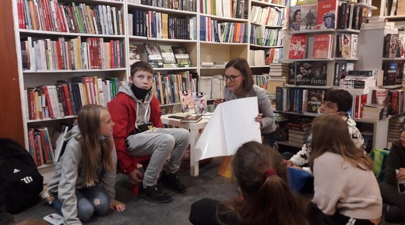 Księgarnia. Przy ścianach białe regały z książkami. W centrum uczniowie siedzą na dywanie. Pani prowadząca zajęcia trzyma w dłoniach blok z białymi kartkami.