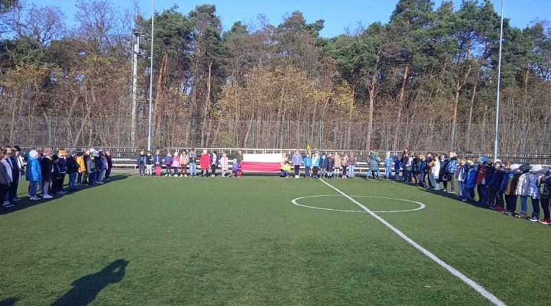 Uczniowie klas 1-3 stoją na boisku szkolnym trzymając flagę Polski