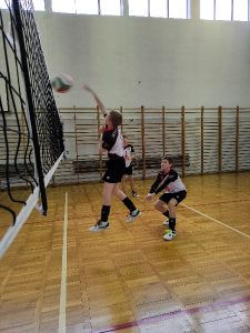 Zdjęcie jest zrobione przy siatce. Siatka znajduje się z lewej strony. W centralnym miejscu zdjęcia zawodnik Tymon jest w wyskoku i atakuje piłkę. Za nim na parkiecie dwóch kolegów z drużyny asekurują atak. Piłka znajduje się nad siatką. Zawodnicy są ubrani w stroje klubowe w kolorach białym i czarnym.