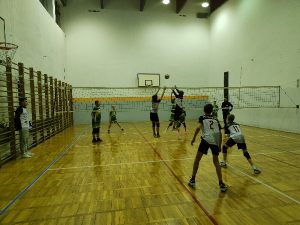 Zdjęcie jest zrobione z linii końcowej boiska. Plecami do aparatu znajdują się zawodnicy UKS Czerwonak. Dwóch nich na środku siatki tworzą blok podwójny. Pozostała dwójka asekuruje blok kolegów. Piłka znajduje się nad ich rękoma. Po drugiej stronie siatki jest przeciwna drużyna z klubu AZS Olsztyn. Mają stroje w kolorach zielonym – białym. Po lewej stronie przy siatce stoi sędzia meczu. Za nim są drabinki gimnastyczne W tle jest ścian.