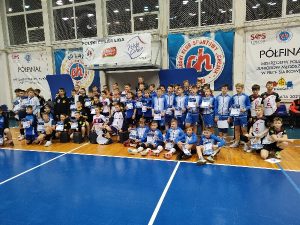 Na środku pozują wszyscy zawodnicy poszczególnych klubów. Zdjęcie zbiorowe. Zaraz od prawej strony są sportowcy UKS Czerwonak. W tle banery reklamowe klubu Chemika Bydgoszcz w kolorze granatowym oraz Półfinał Mistrzostw Polski Juniorów Młodszych w Piłce Siatkowej koloru białego. U góry okna hali, a na dole widać parkiet w kolorze granatowym.