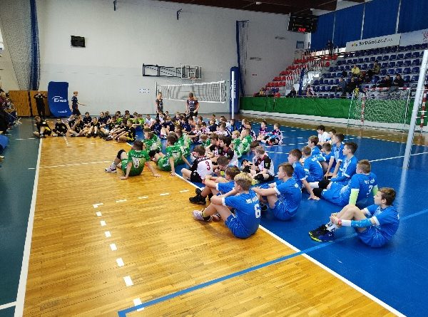 Otwarcie zawodów. Wszyscy zawodnicy siedzą zespołami na parkiecie hali. Podłoga jest wielobarwna z różnymi pomalowanymi liniami do zespołowych gier sportowych. W tle znajduje się rozłożona siatka do gry oraz trybuny.