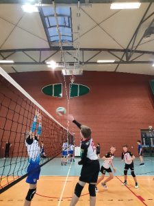 Zdjęcie wykonano przy siatce. Na prawo od siatki zawodnik Aleksander z klubu UKS Czerwonak wykonuje atak prawą ręką. Po drugiej stronie siatki ubrany w stój sportowy koloru biało – granatowego zawodnik drużyny przeciwnej wykonuje blok pojedynczy. Piłka znajduje się pomiędzy zawodnikami nad siatką. Przy zawodniku atakującym znajduje się dwóch kolegów z drużyny i asekurują. W tle znajduje się kolejny mecz innych drużyn oraz ściana hali w kolorze brązowym. Przy górnej krawędzi zdjęcia znajduje się świetlik (okno)