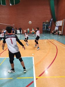 Na zdjęciu widać czterech zawodników klubu UKS Czerwonak ustawieni w przyjęciu piłki. Jeden z nich stoi plecami do aparatu, ma na stroju sportowym w kolorze biało-czarnym napis „UKS CZERWONAK” oraz numer 1. W głębi zdjęcia znajdują się pozostali zawodnicy zespołu i jeden z nich odbija piłkę sposobem oburącz dolnym. Pozostali ustawieni są na swoich pozycjach przy siatce i na prawym skrzydle. W tle widać jeszcze jeden mecz oraz ścianę koloru brązowego. Parkiet hali jest dwubarwny – kremowy i szarozielony. Ma namalowane linie czerwone i białe