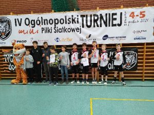 Zdjęcie przedstawia dwa zespoły ustawione w rzędzie trzymające dyplomy za udział w turnieju. Jedna z drużyn to UKS Czerwonak stojąca po prawej stronie. Na drugiej stronie rzędu znajduje się maskotka organizatora – tygrys klubu UKS 9 Leszno. Zawodnicy i maskotka za plecami mają drabinki gimnastyczne. Nad ich głowami umieszczony jest bardzo duży baner z napisem „IV Ogólnopolski Turniej Mini Piłki Siatkowej – 4/5 grudnia 2021 r” oraz logo klubu (twarz tygrysa na czarnym tle). Na dole zdjęcia widać podłogę hali w kolorze szarozielonym.