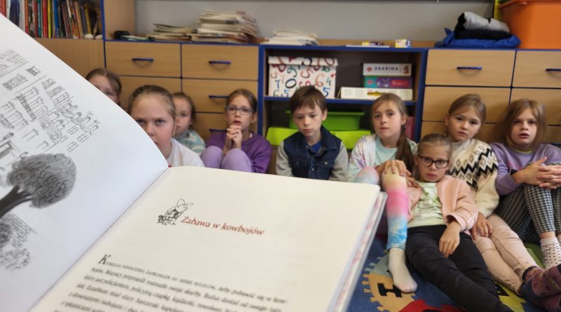 Książką, a w tle zasłuchani uczniowie siedzący na dywanie