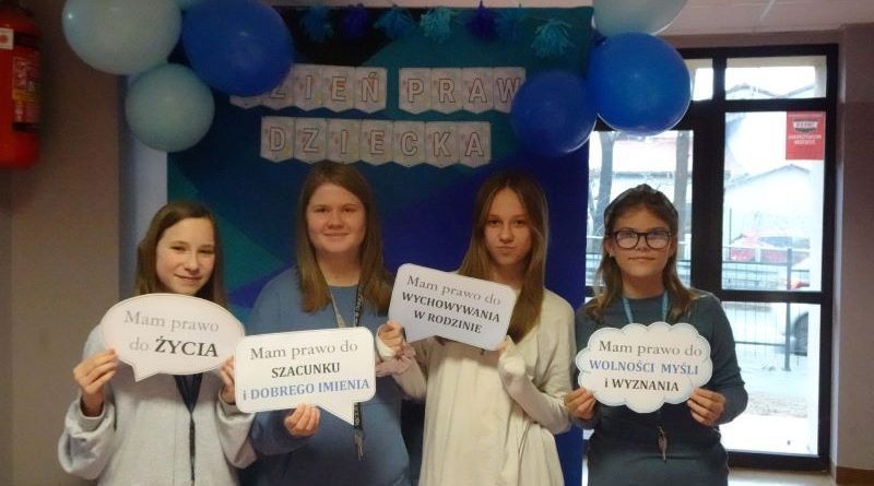 Zdjęcie nr 3 - cztery dziewczynki z klasy 6 stoją na tle niebieskiej ścianki z napisem Dzień Praw Dziecka ozdobionej niebieskimi balonami, dwie dziewczynki mają jasne, kremowe bluzy a dwie mają niebieskie bluzy, wszystkie trzymają chmurki z napisami mam prawo do życia, szacunku i dobrego imienia, do wychowania w rodzinie, do wolności myśli i wyznania; w tle widać duże okno i drzwi.