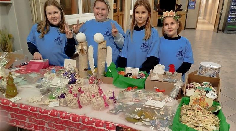 Na zdjęciu widać 4 uczennice – wolontariuszki stojące w holu szkoły, w równych błękitnych koszulkach. Przed nimi znajduje się stół nakryty wzorzystym, czerwonym obrusem. Na nim ułożone są różne przedmioty na sprzedaż: dwa duże anioły z drewna, bombki , gwiazdki, choinki z makramy, ozdoby z masy solnej , aniołki i czapeczki ze sznurka.