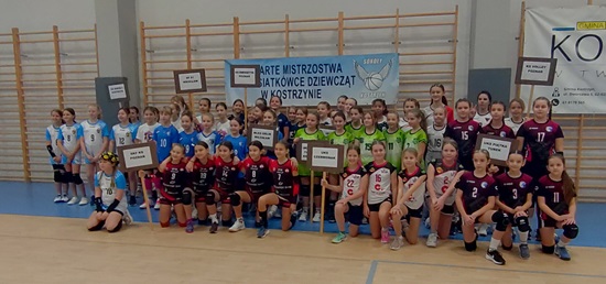 To zdjęcie przedstawia wszystkie zawodniczki, które stawiły się na Otwartym Turnieju Minisiatkówki w Kostrzynie. Dziewczęta pozują na tle błękitnego baneru. Część klęczy a część stoi. Ubrane są w stroje klubowe o wielobarwnych kolorach. Dziewczęta z UKS Czerwonak mają na sobie stroje klubowe w kolorach biało-czarno-czerwonym. W tle widać drabinki gimnastyczne.