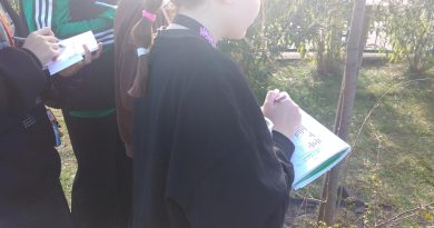 Czwórka dziewcząt na terenie szkolnym obserwuje porosty na drzewie, trzymając w ręce zeszyt z kartą pracy i długopis.