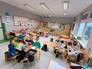 Dzieci siedzą w ławkach w grupach kilkuosobowych. Wszyscy patrzą w obiektyw aparatu.