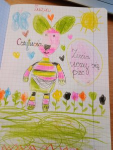 Na kartce narysowana jest maskotka i podpisana imieniem Czytusia. Ma ona kolorowe ubranie a wokół niej rosną kwiatki. Po prawej stronie znajduje się napis : Zuzia uczy się piec. 