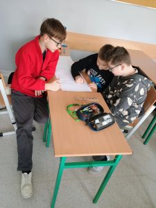 Trzech chłopców siedzi w ławce. Są pochyleni nad kartką papieru. 