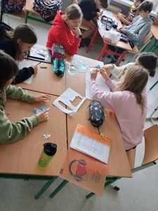 y stolikach, w grupach siedzą uczniowie i wspólnie pracują nad plakatem.