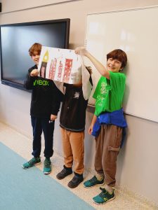 Przed tablicą stoi trzech chłopców. Podnoszą do góry zrobiony przez siebie plakat.