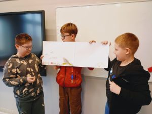 rzech uśmiechniętych chłopców prezentuje swój plakat. Stoją oni przed monitorem interaktywnym.
