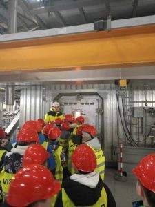 Widać na zdjęciu grupę uczniów ubranych w czerwone kaski i kamizelki odblaskowe stojących w rzędzie przed wielkim kotłem, gdzie odbywa się proces spalania odpadów. Kozioł jest stalowy i posiada małą szybę, przez którą można zobaczyć płomienie wytwarzające temperaturę około 700 stopni Celcjusza. Obok okienka stoi przewodnik i nadzoruje pokaz