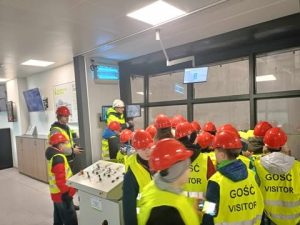 Uczniowie stoją ubrani w strój ochronny w centrali zakładu. Obserwują pracę operatora suwnicy i chwytaka. W tle widać bardzo duże okna a za nimi śmieci i chwytak. Na jasnej ścianie widać kilka monitorów z danymi.