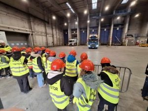 Na zdjęciu znajdują się uczniowie przy barierce ochronnej. Są dodatkowo ubrani w czerwone kaski i kamizelki odblaskowe. Obserwują plac rozładunkowy. Widać na końcu placu pięć dużych niebieskich bram przez, które są zrzucane śmieci do bunkra na odpady. Przy dwóch bramach stoją śmieciarki.