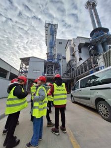 Na tym zdjęciu widać zakład z zewnątrz. Uczniowie przypatrują się całemu budynkowi wraz z instalacją ciepłowiniczą i wodną. Po prawej stronie widać dwa duże kominy oraz samochód. Nad zakładem widoczne jest niebo wraz z chmurami. Uczniowie są ubrani dodatkowo w kaski czerwone i kamizelki odblaskowe.