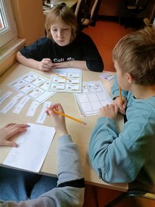 trzej chłopcy siedzący wokół stołu, trzymają w rękach ołówki, na twarzy jednego z nich widoczne skupienie, pracują pochyleni nad kartkami.