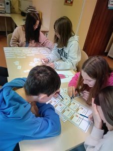 ujęcie z góry. Przy stole umieszczonym dalej siedzą dwie dziewczyny, pracują, na stole rozłożone kartki. Przy drugim stole, bliżej oglądającego, widać trzy osoby - chłopca i dwie dziewczyny. Pochyleni są nad kartkami z rysunkami. Z gestów widać, że jedna z dziewcząt śmieje się.