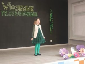 Na scenie stoi dziewczynka w zielonej sukience i białym sweterku. Jej blond włosy opadają na ramiona.