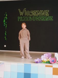 Na scenie stoi dziewczynka w brązowych spodniach i bluzie. Ręce trzyma w kieszeniach.