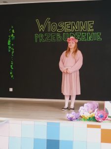 Na scenie stoi dziewczynka, która na głowie ma rózowy wianek zrobiony z kwiatków. Ubrana jest w sukienkę.