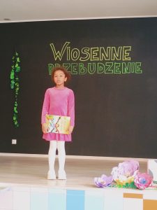 Na scenie stoi dziewczynka, która trzyma przed sobą książeczkę. Ubrana jest w różową sukienkę.