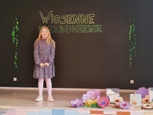 Na scenie stoi dziewczynka z rękoma skrżyżowanymi przed sobą. Ubrana jest w sukienkę oraz białe rajstopy. Włosy ma rozpuszczone i opadające na ramiona.