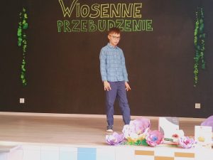 Na scenie stoi chłopiec w niebieskiej koszuli i jeansowych spodniach.