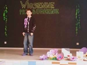 Na scenie stoi chłopiec, który trzyma ręce przed sobą. Po jego lewej stronie ułożone są na podłodze papierowe kwiaty.