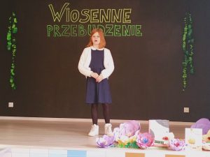 Na scenie stoi dziewczynka w granatowej sukience i białym sweterku.