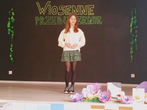 Na scenie stoi dziewczynka . Ubrana jest w krótką, zieloną spódniczkę oraz biała bluzeczkę.