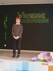 Na scenie stoi chłopiec z rękoma skrzyżowanymi z przodu.