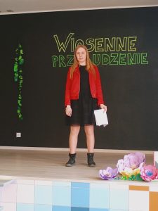 Na scenie stoi wysoka dziewczynka, która w lewej ręce trzyma kartkę. Ubrana jest w czarną sukienkę i czerwoną kurteczkę. 