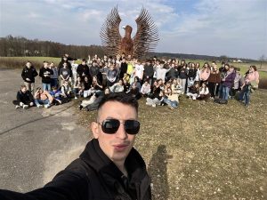 Na pierwszym planie stojący przodem do aparatu mężczyzna ubrany w czarną kurtkę. Nosi ciemne okulary przeciwsłoneczne. Z tyłu za nim bardzo liczna grupa na tle dużej figury orła.