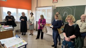 Grupa dyrektorów i nauczycieli w sali lekcyjnej. W tle tablica interaktywna.