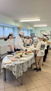 Uczniowie w fartuszkach i czapkach kucharskich podczas warsztatów kulinarnych. 