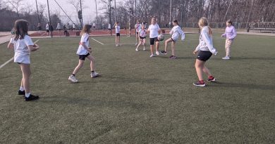 Na zdjęciu widzimy uczenice klas 5 walczące o piłkę w trakcie meczu piłki nożnej. Med odbywa się na boisku ze sztuczną nawierzchnią, zawodniczki ubrane w stroje sportowe, oznaczone są szarfami w kolorach żółtym o czerwonym.