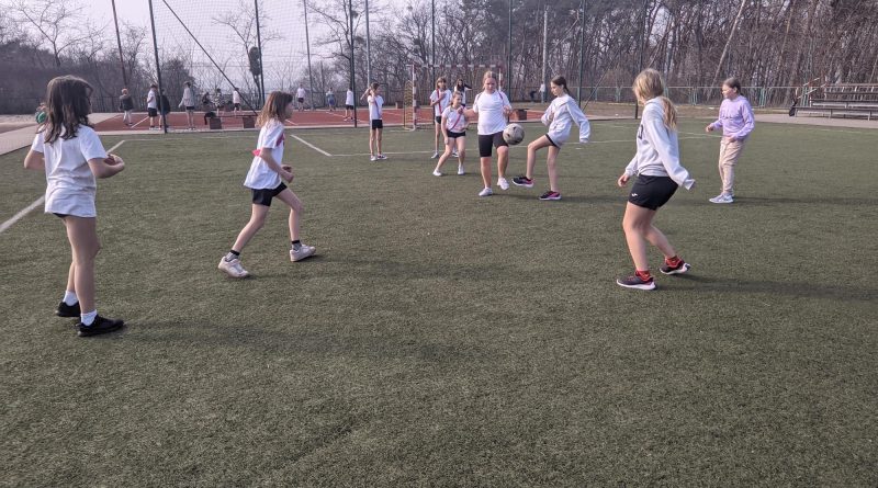 Na zdjęciu widzimy uczenice klas 5 walczące o piłkę w trakcie meczu piłki nożnej. Med odbywa się na boisku ze sztuczną nawierzchnią, zawodniczki ubrane w stroje sportowe, oznaczone są szarfami w kolorach żółtym o czerwonym.