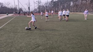 Na zdjęciu widzimy uczenice klas 5 walczące o piłkę w trakcie meczu piłki nożnej. Med odbywa się na boisku ze sztuczną nawierzchnią, zawodniczki ubrane w stroje sportowe, oznaczone są szarfami w kolorach żółtym o czerwonym.