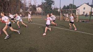 Na zdjęciu widzimy uczenice klas 5 walczące o piłkę w trakcie meczu piłki nożnej. Med odbywa się na boisku ze sztuczną nawierzchnią, zawodniczki ubrane w stroje sportowe, oznaczone są szarfami w kolorach żółtym o czerwonym.