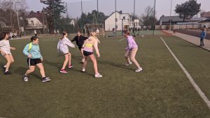 Na zdjęciu widzimy uczenice klas 5 walczące o piłkę w trakcie meczu piłki nożnej. Med odbywa się na boisku ze sztuczną nawierzchnią, zawodniczki ubrane w stroje sportowe, oznaczone są szarfami w kolorach żółtym o czerwonym.